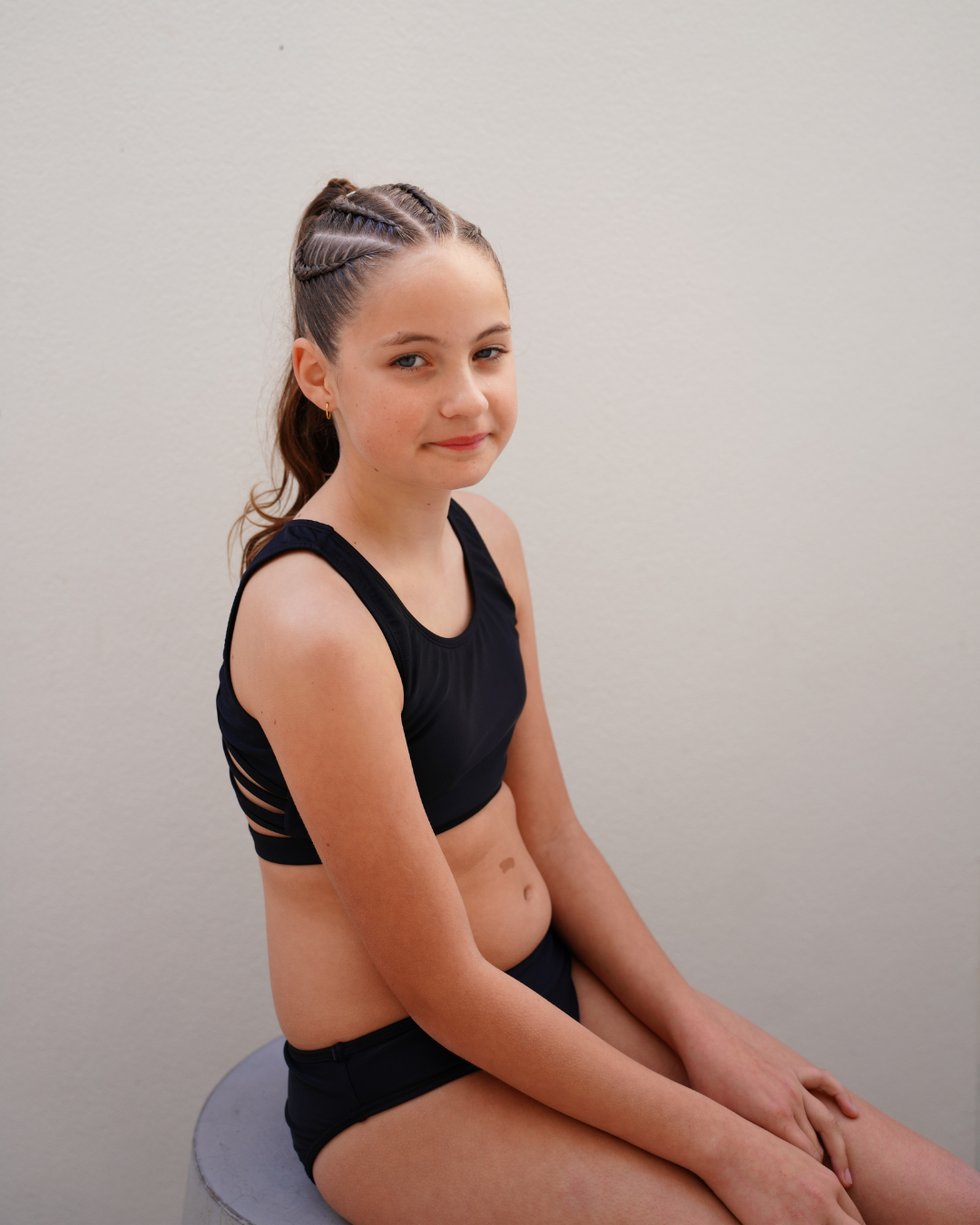 A young girl with braided hair sits on a stool wearing a black girls tween bikini and matching high-waisted briefs against a plain light gray wall.