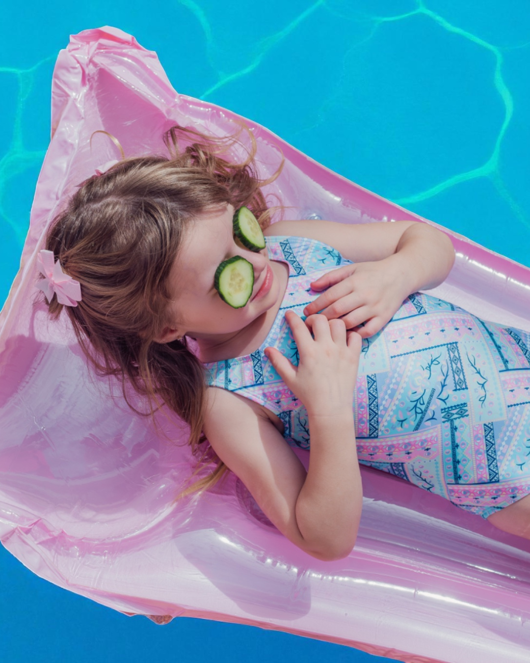 Child lying on a pink inflatable raft in a pool with cucumbers on their eyes.
