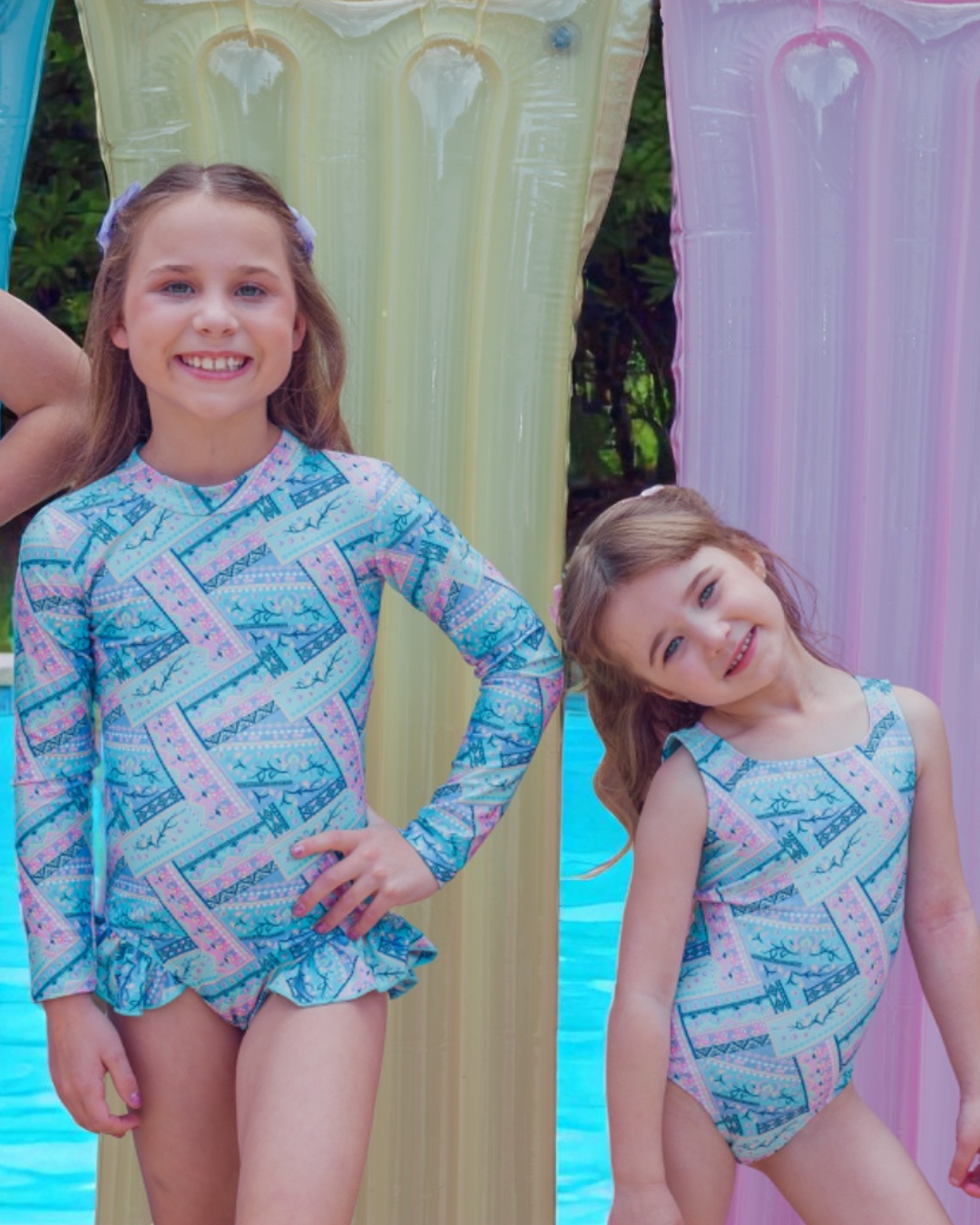 Two children wearing matching girls swimsuits by a poolside with colorful curtains.