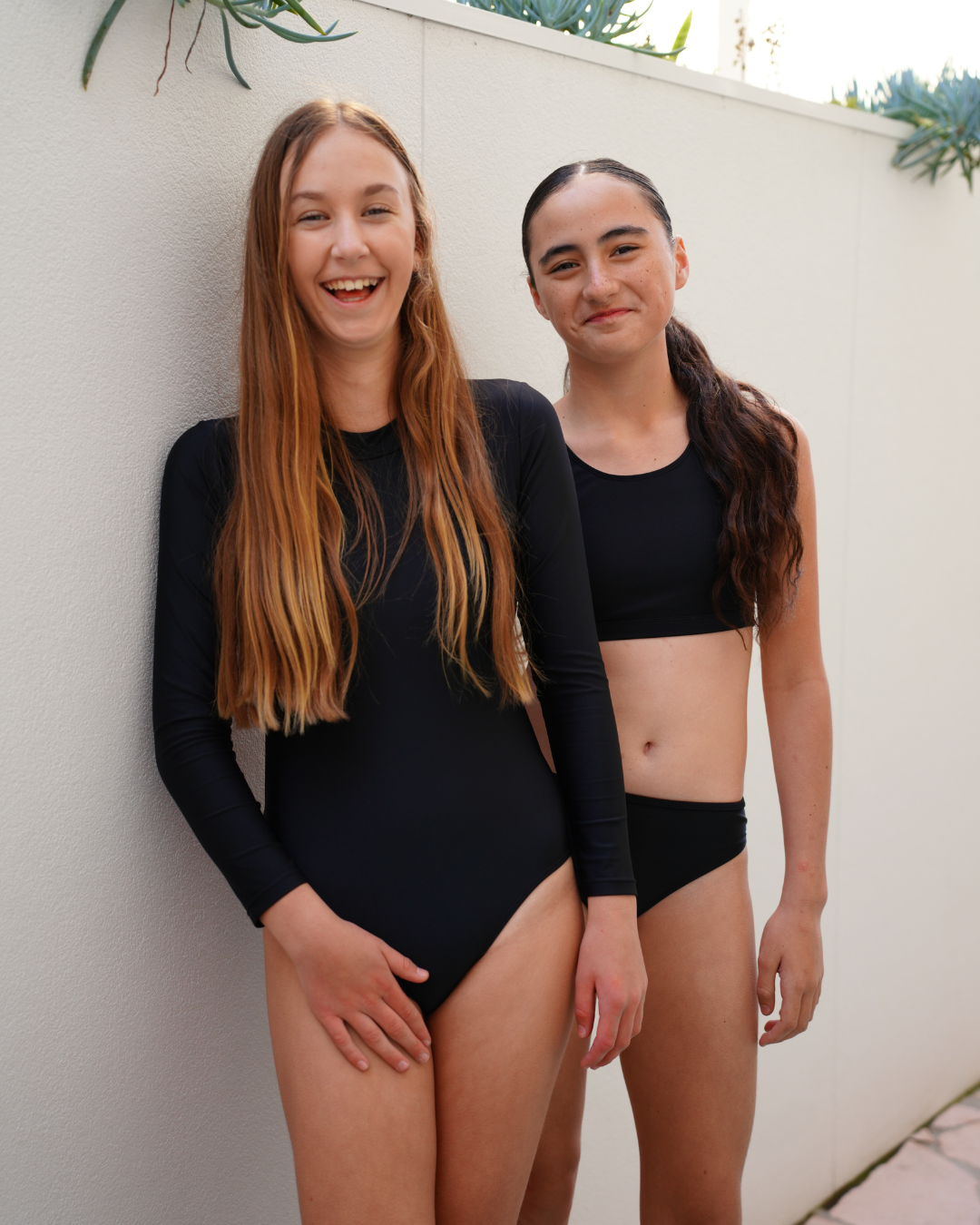 Two young female swimmers posing outdoors against a white stucco wall. The girl on the left wears a black long-sleeve one-piece girls swimsuit, and the girl on the right wears a black two-piece swimsuit consisting of a crop top and bikini bottoms.