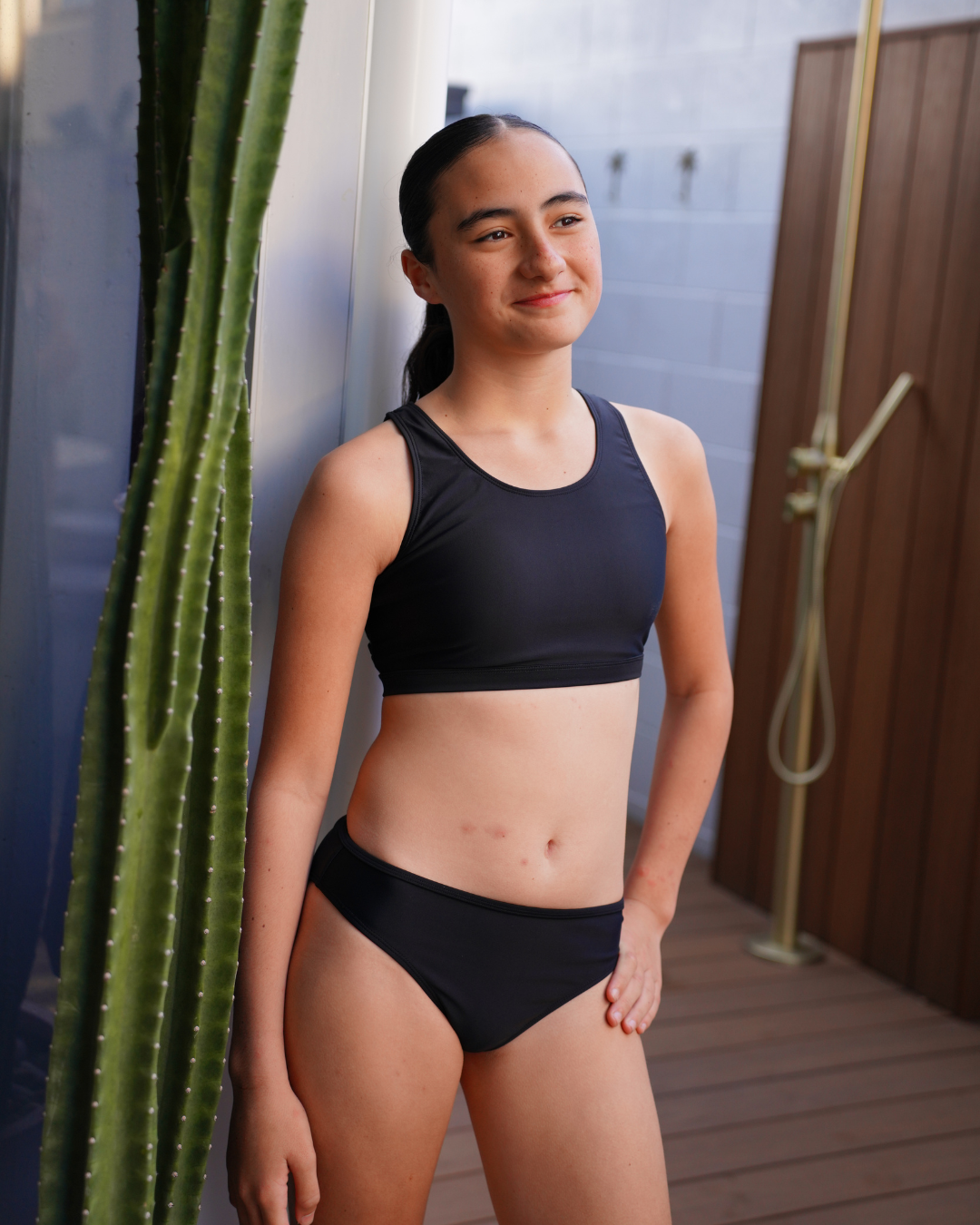 A woman wearing a black two-piece girls swimsuit stands on a wooden deck next to a tall green cactus, with a modern outdoor shower visible in the background.