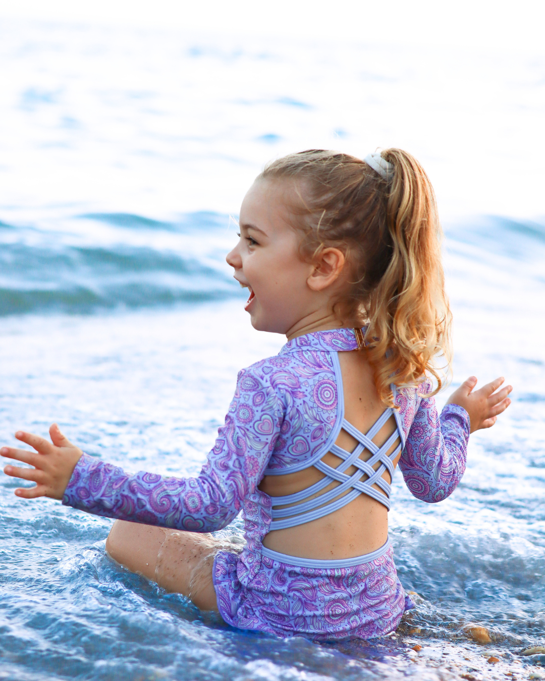 A young girl with a ponytail sits in shallow ocean water wearing a purple and lavender patterned long-sleeve girls bathers with crisscross back straps and matching swim bottoms.