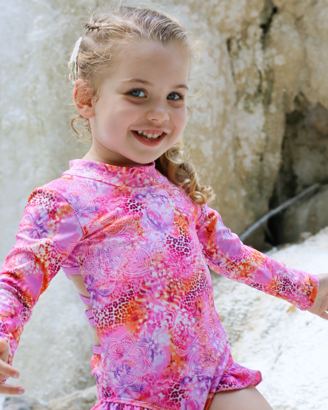 A young girl smiling and wearing a pink long-sleeve swimsuit with a colorful abstract pattern, set against a natural rocky background.