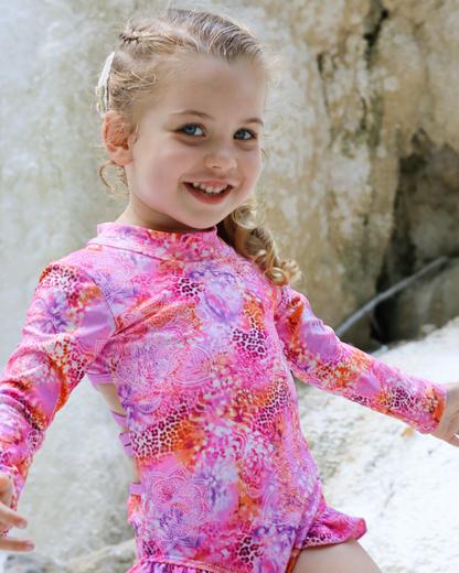 A young girl smiling and wearing a pink long-sleeve swimsuit with a colorful abstract pattern, set against a natural rocky background.