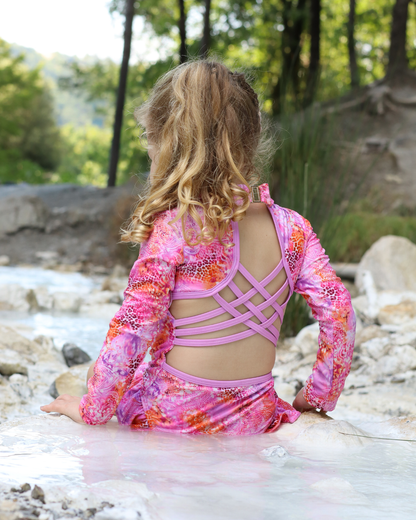 A young girl with blonde hair is seen from behind, sitting in shallow water outdoors. She is wearing a pink long-sleeve swimsuit with a vibrant floral pattern in pink, orange, and purple.