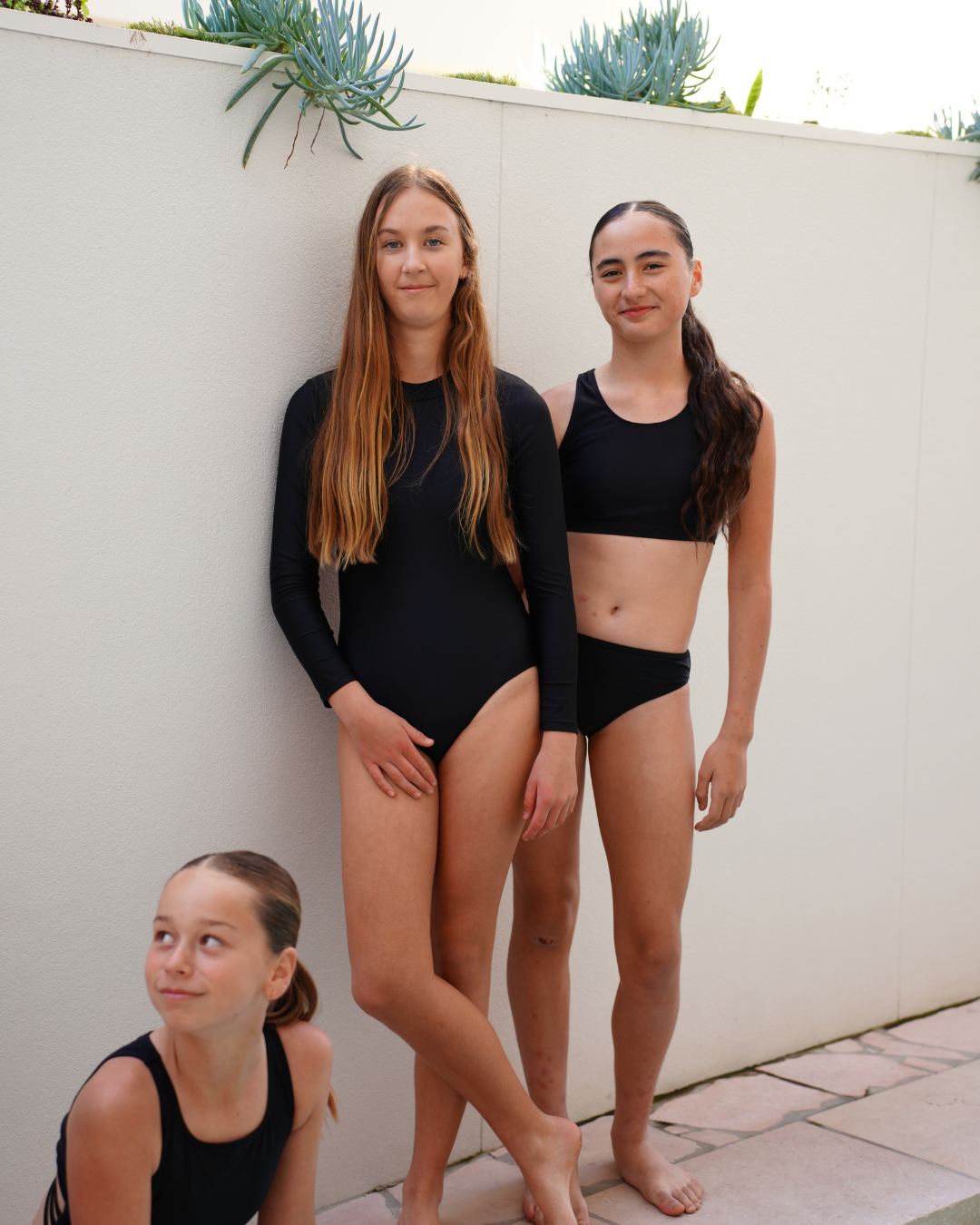 Three young female swimmers posing in black athletic swimwear against a white wall.