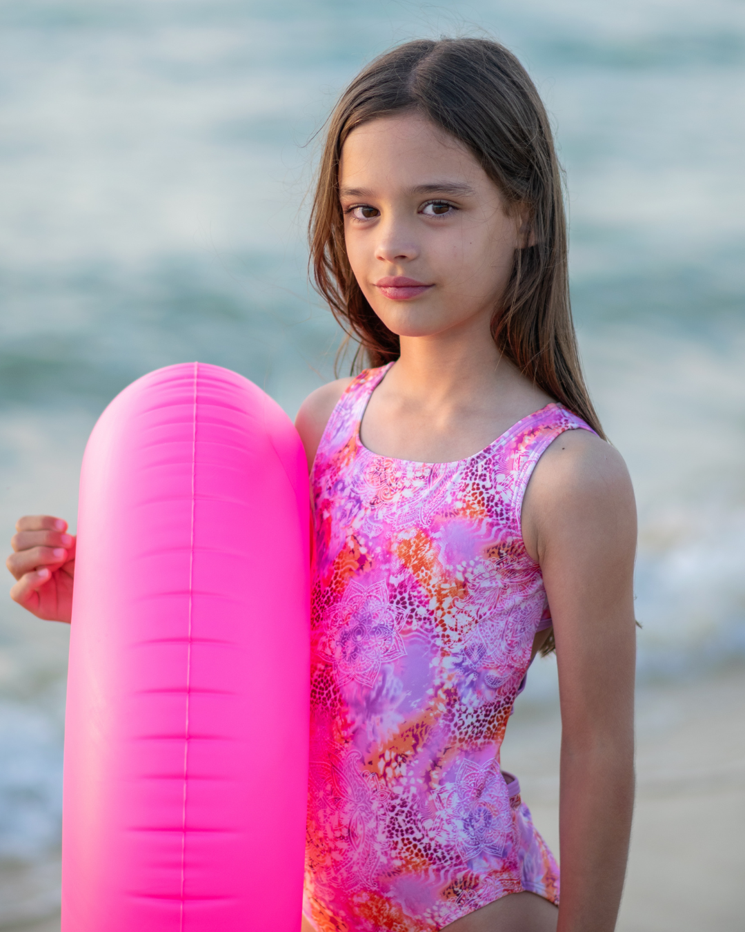 Young girl holding a pink inflatable ring by the water and wearing a one piece girls swimsuit