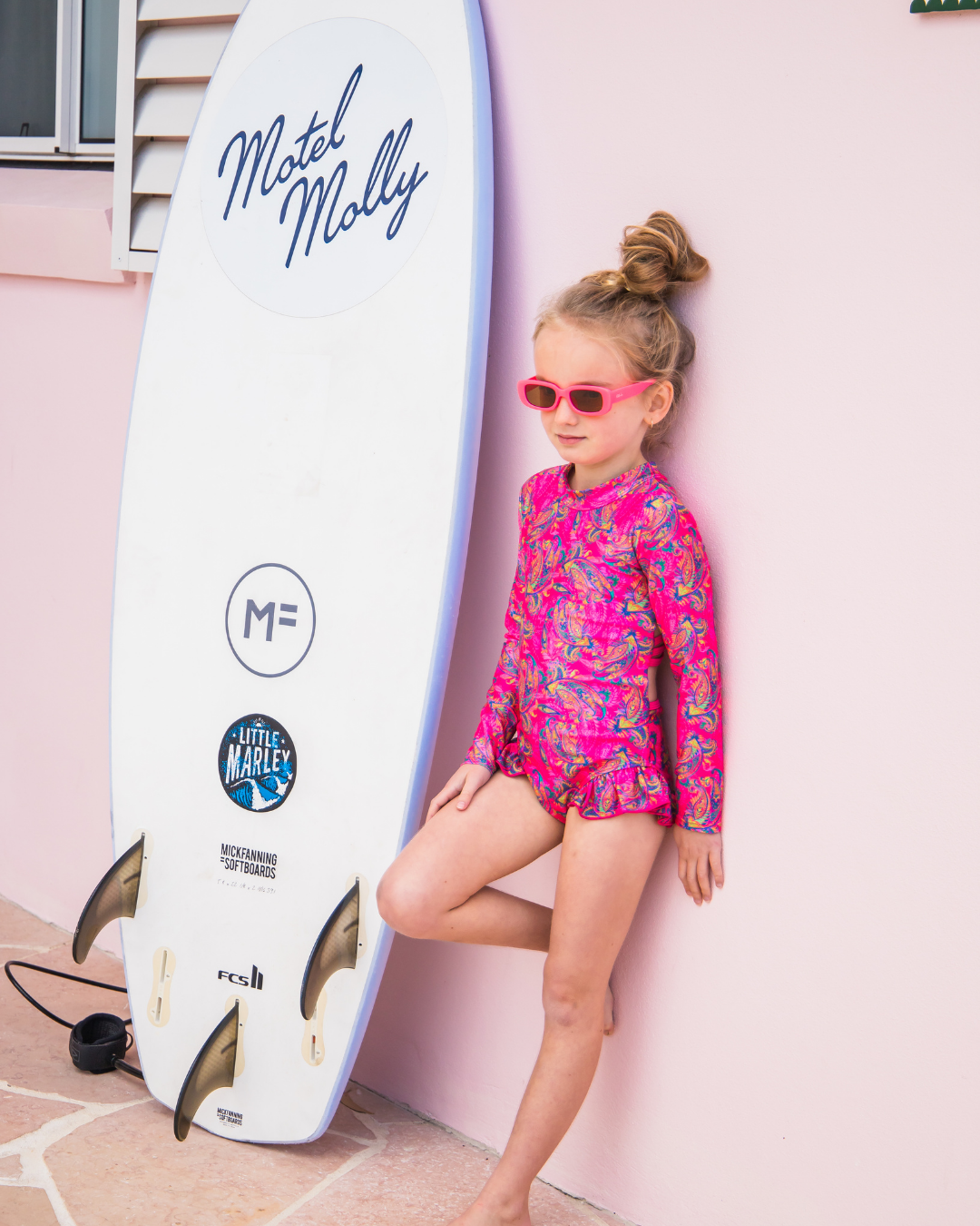 Child in a pink teen swimwear standing next to a Motel Molly surfboard.