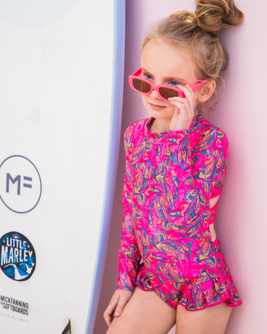 Child wearing a bright pink and blue patterned teen swimwear with sunglasses, standing next to a surfboard.