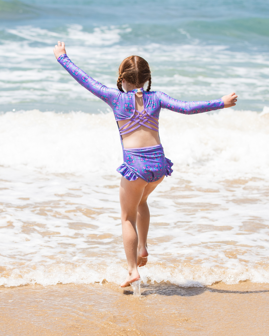 Child in a purple kids swimwear standing on a beach with arms outstretched.