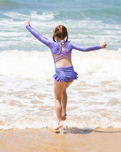 Child in a purple kids swimwear standing on a beach with arms outstretched.