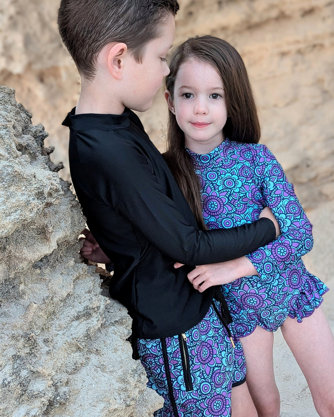 Two children in matching swimsuits standing close to each other against a natural stone wall.