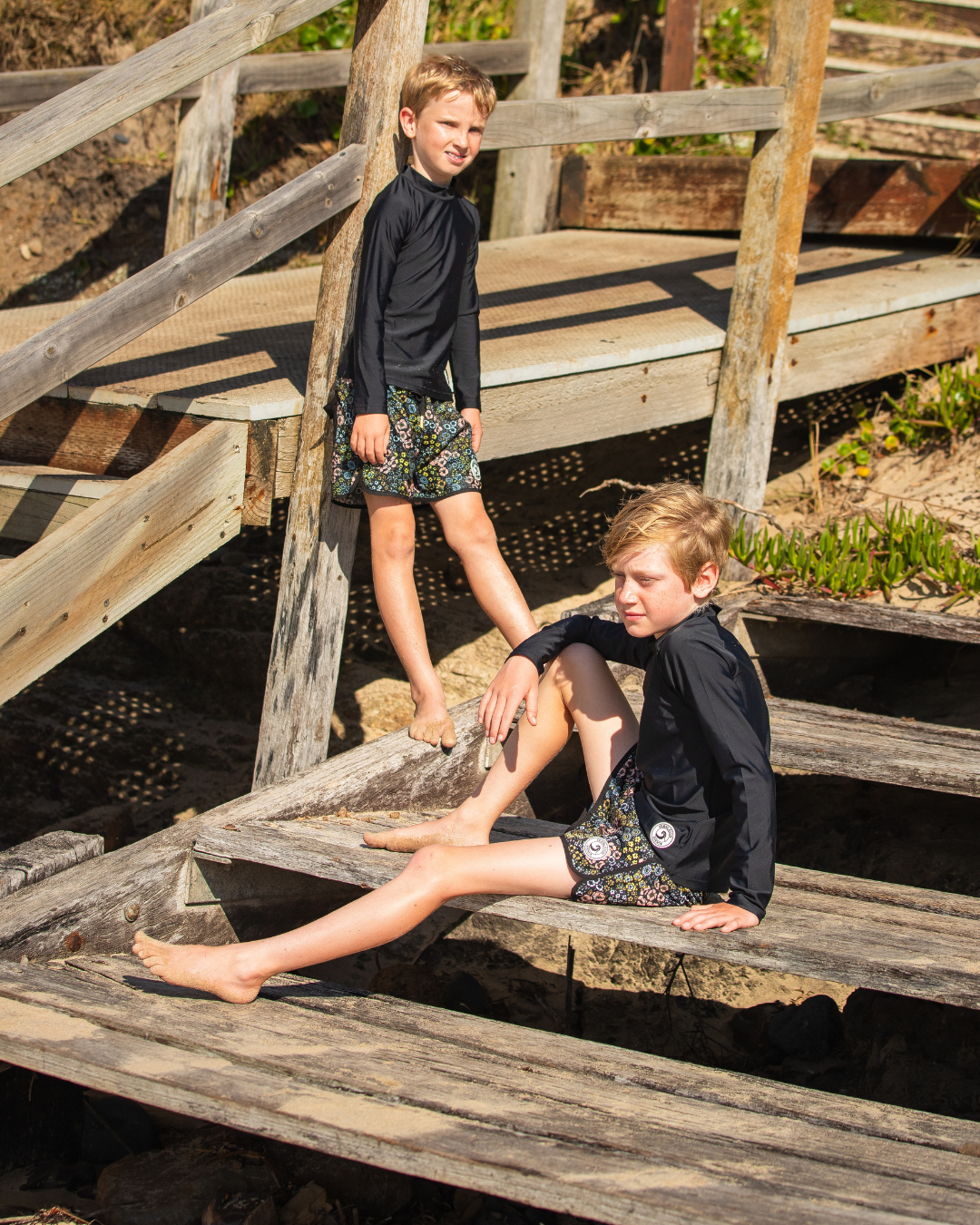 Two children wearing boys board shorts sitting on a wooden staircase outdoors