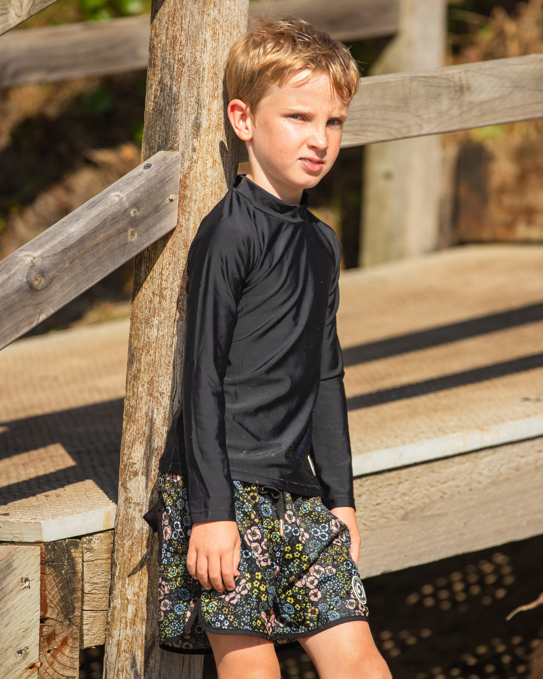 Young boy wearing boys swimwear standing against a wooden fence.
