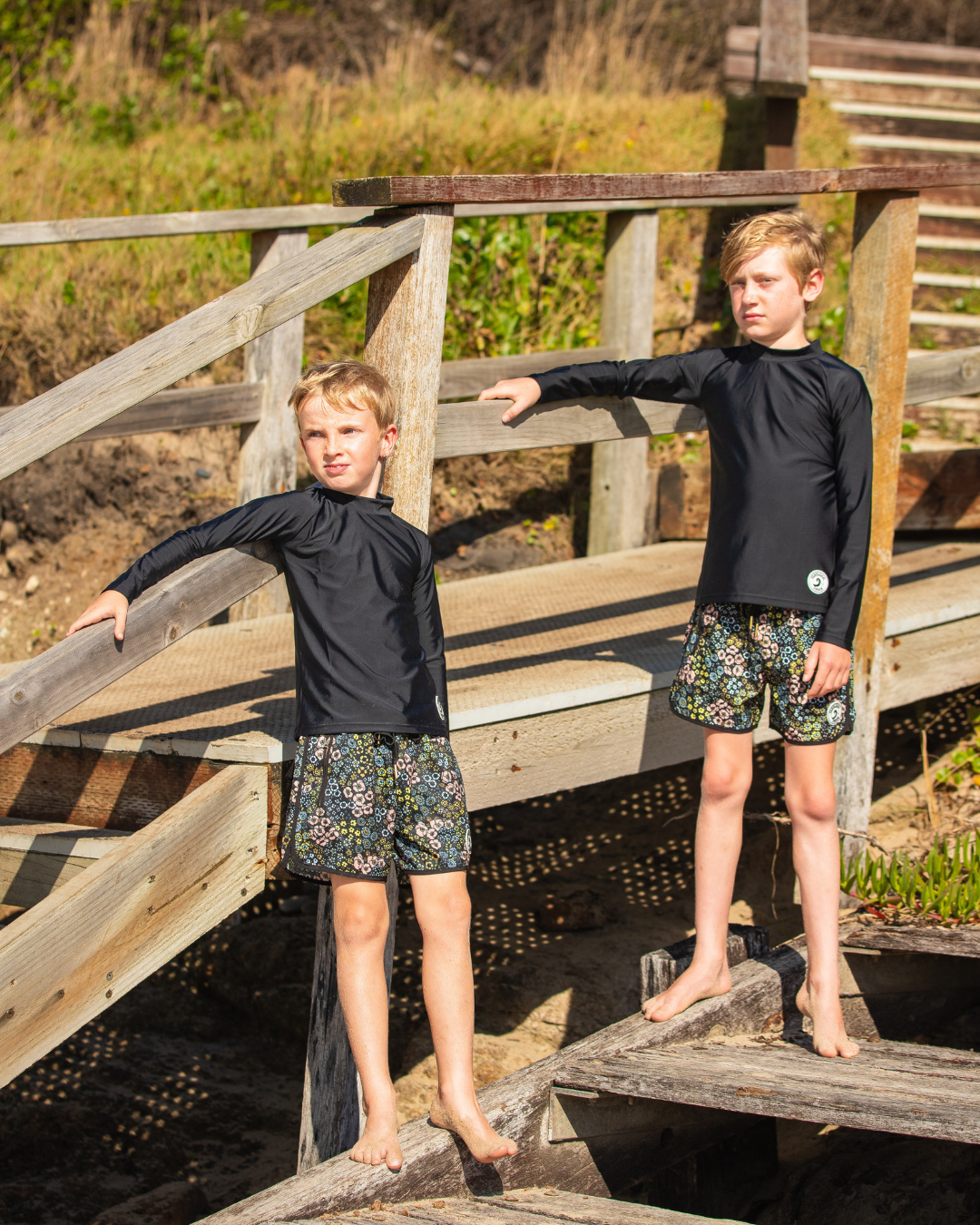 Two children wearing boys swimwear  standing on a wooden bridge.