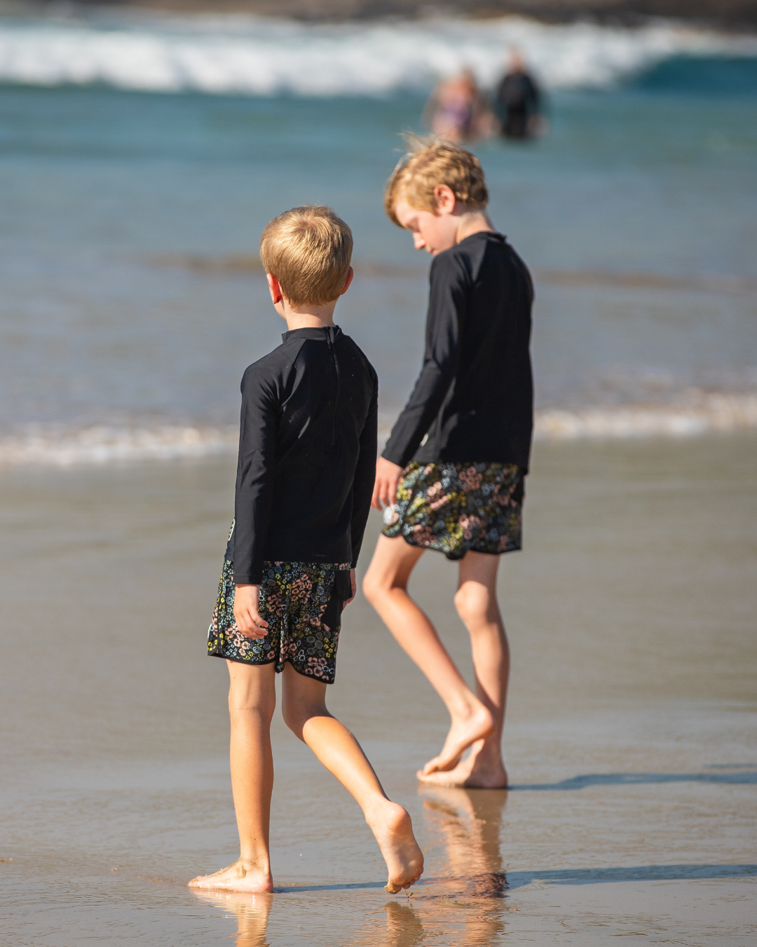 Two children walking on a beach wearing boys swimwear with ocean waves in the background