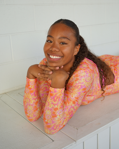 A smiling young woman with dark curly hair tied back lies on her stomach on a white tiled surface, wearing a long-sleeve teen swimsuit with a vibrant orange, pink, and yellow floral pattern.