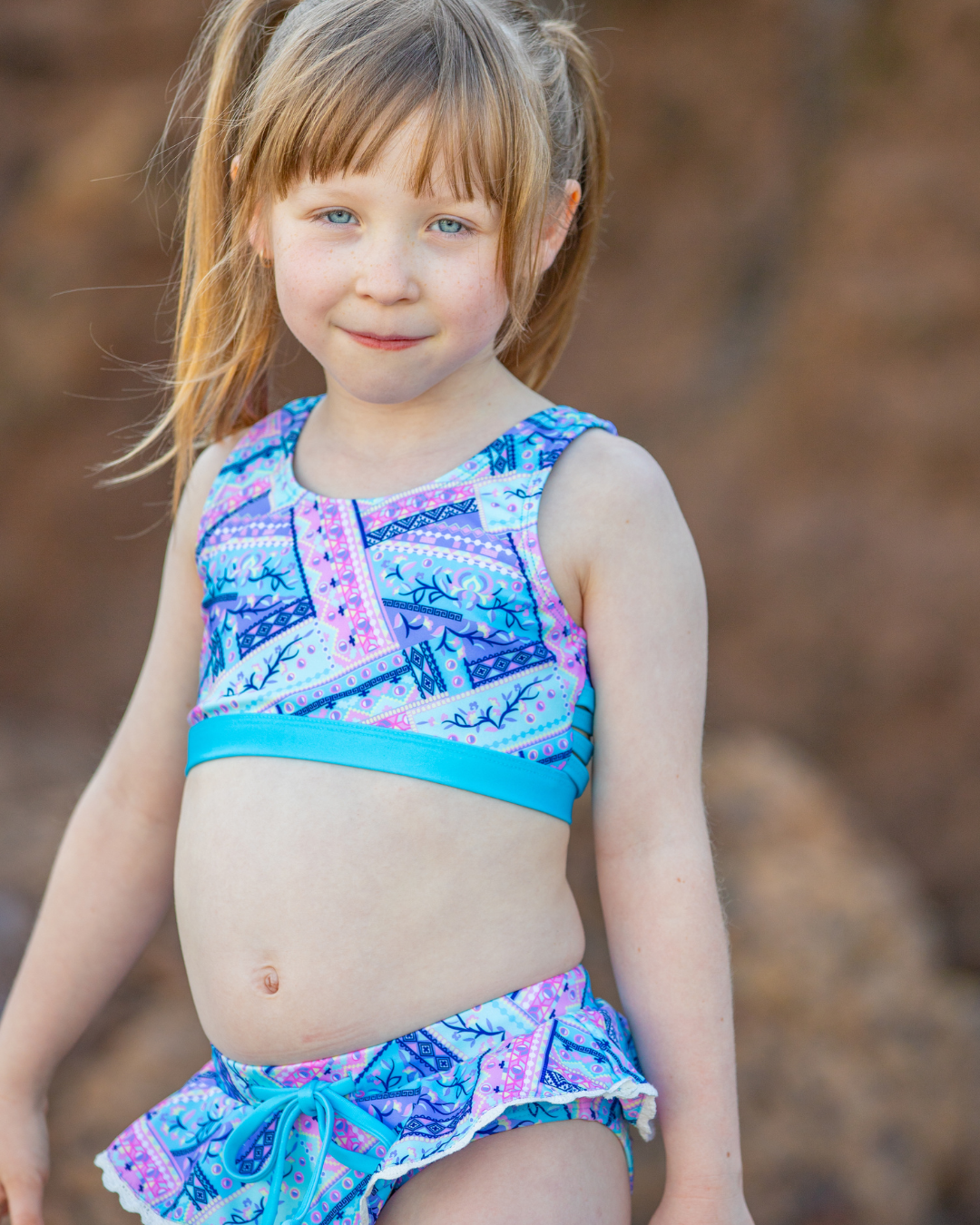 Young girl wearing a colorful tween bikini swimsuit with a patterned top and matching bottoms against a natural background.