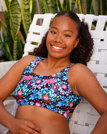 A smiling woman wearing a colorful floral teen bikini sits on a white woven lounge chair with green plants in the background.