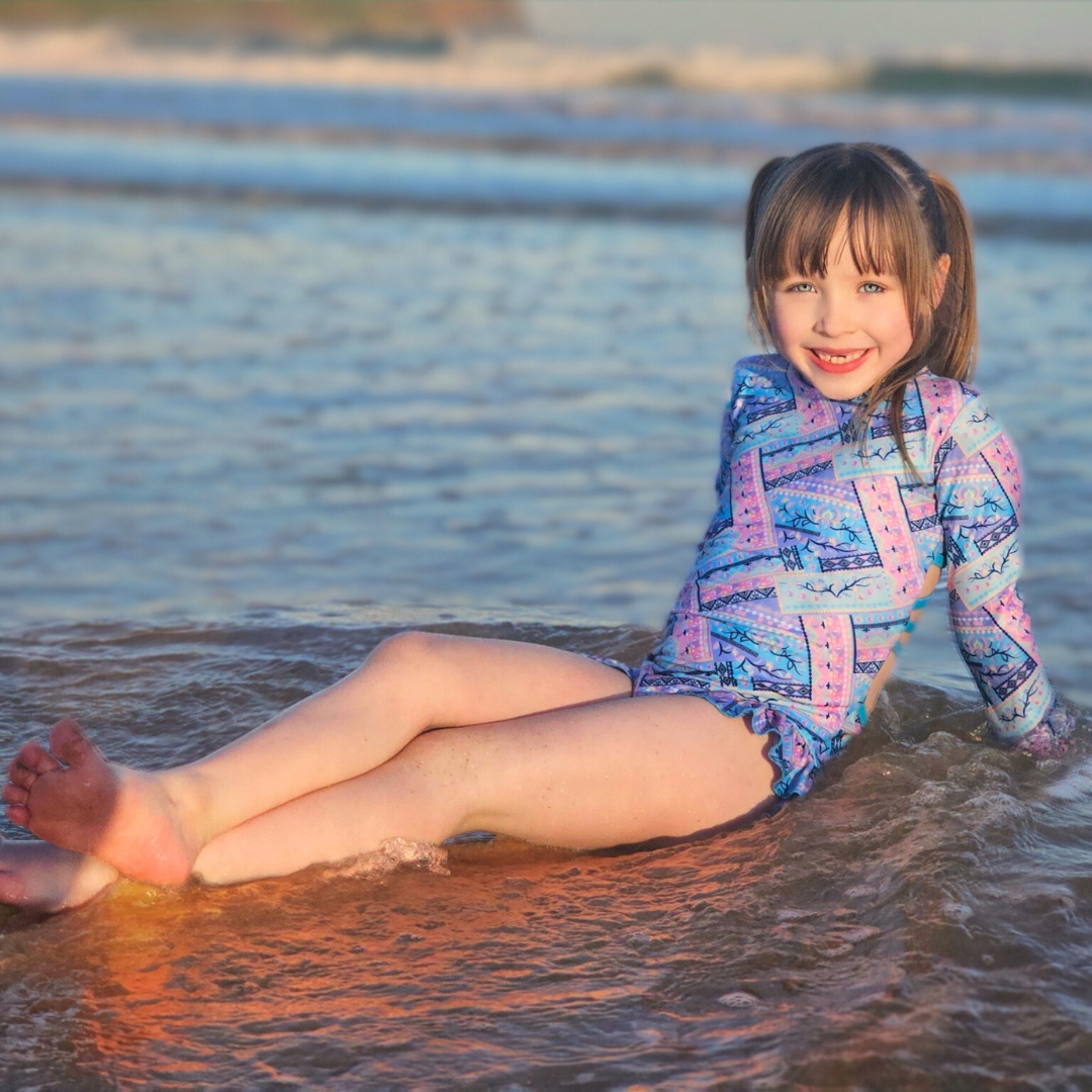 Child in a colorful teen swimwear sitting in shallow water at the beach.
